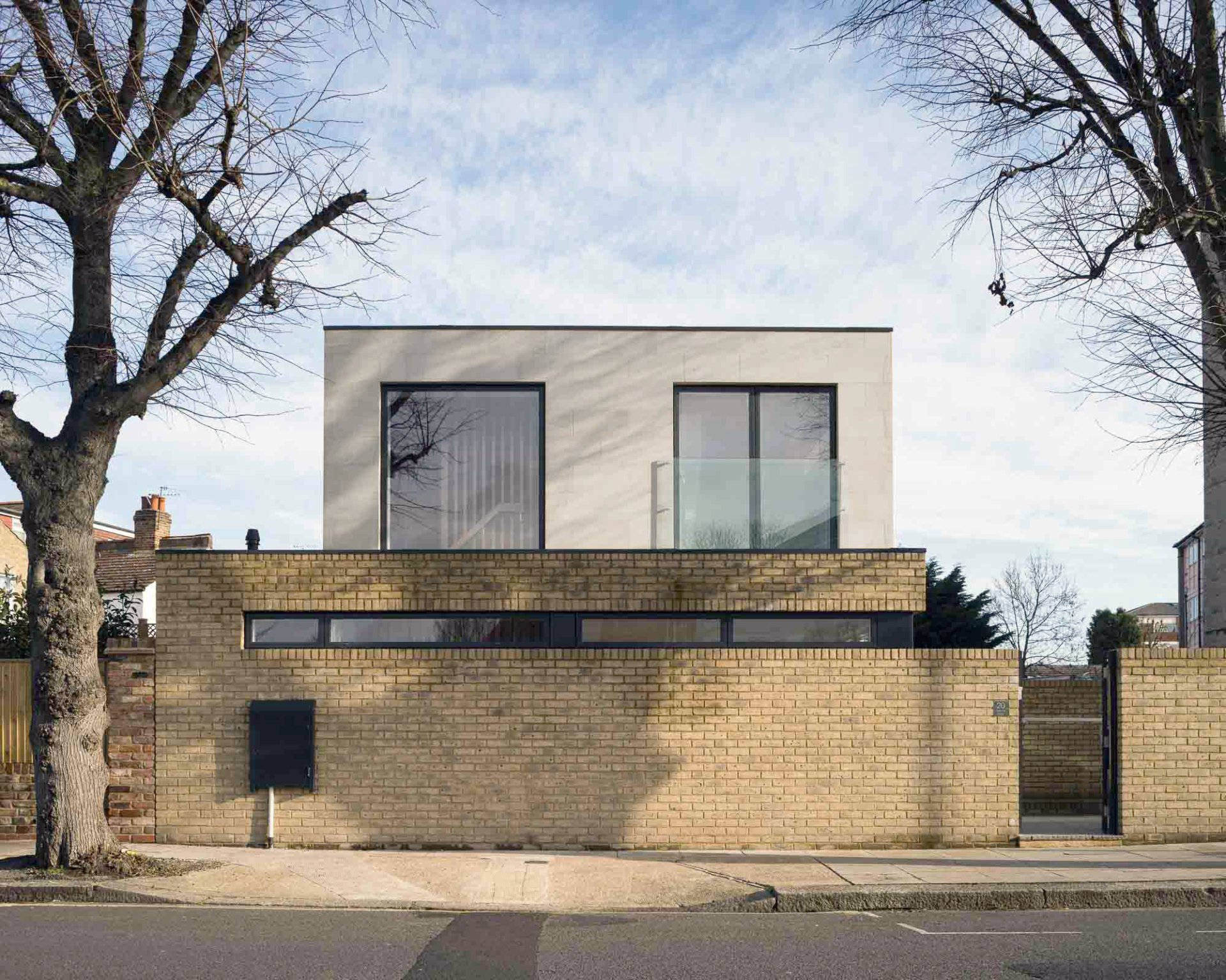 Nowoczesny, dwupiętrowy dom z jasną fasadą na ceglanym murze, widok od strony ulicy. Drzewa otaczają budynek pod błękitnym niebem.