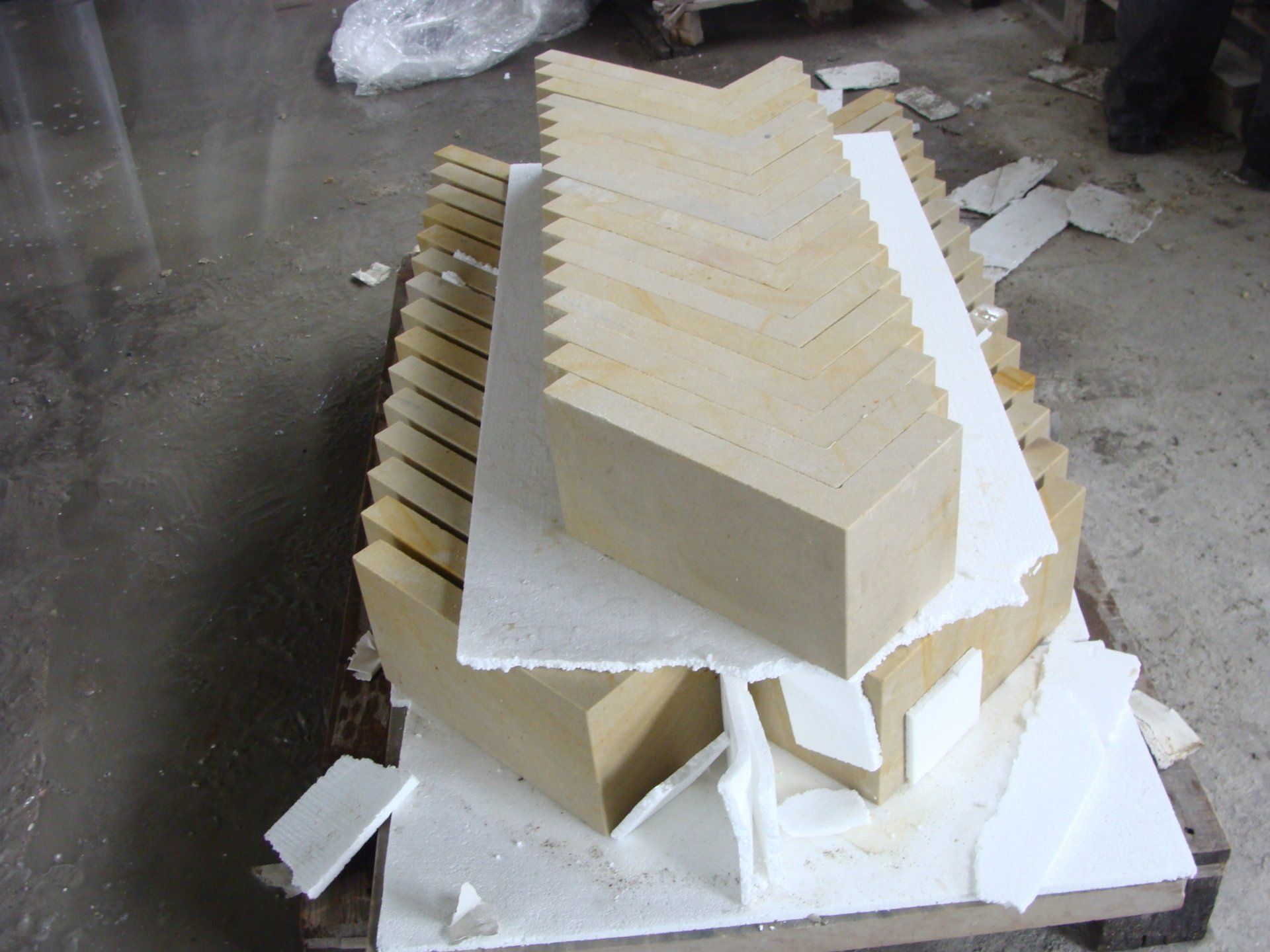 Kawałki cegły w kolorze jasnobrązowym ułożone we wzór jodełki, na białym materiale opakowaniowym, spoczywające na drewnianej palecie w warsztacie.