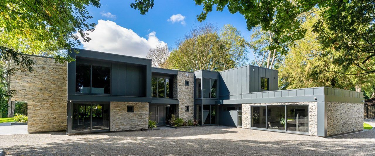 Nowoczesny dom z kamienną fasadą i ciemnoszarą elewacją, dużymi oknami i otaczającymi drzewami.