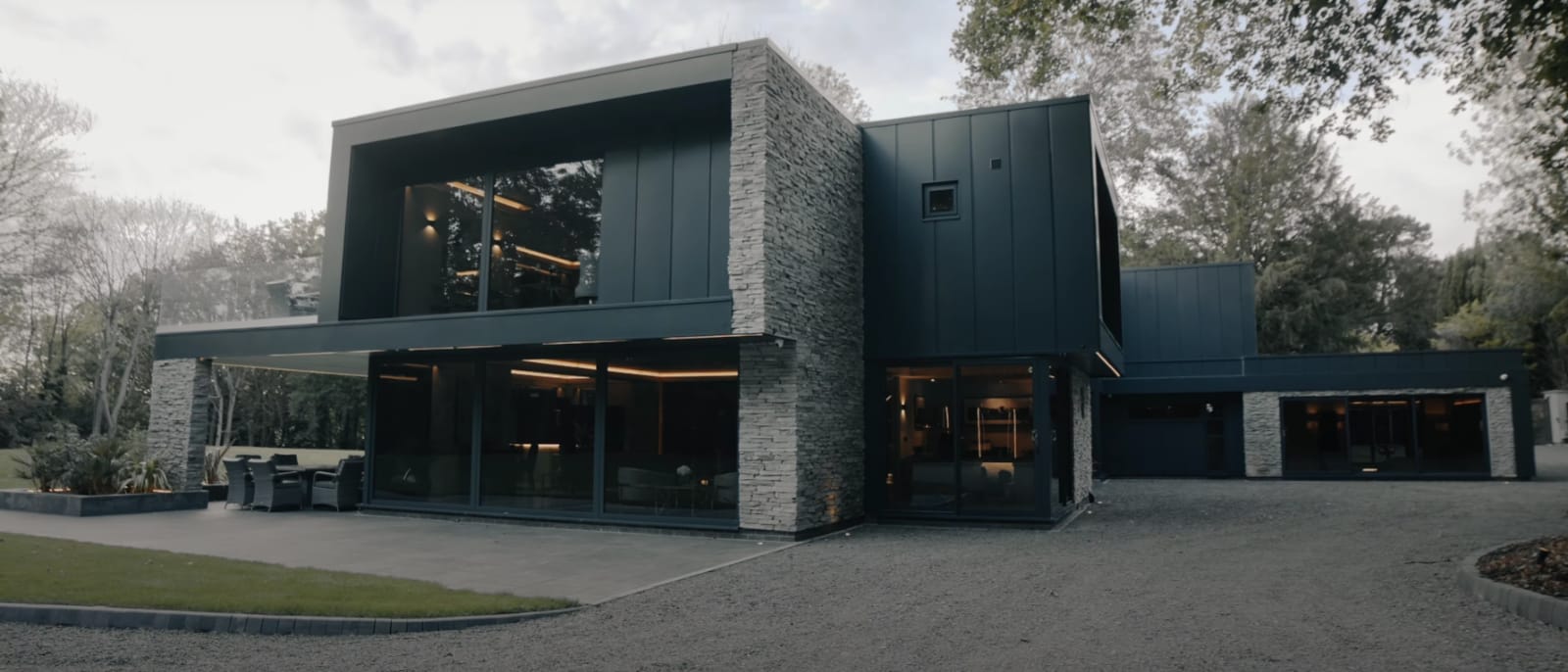 Nowoczesny, dwupiętrowy dom z ciemną elewacją, kamiennymi akcentami i długim podjazdem. Położony wśród drzew i zachmurzonego nieba.