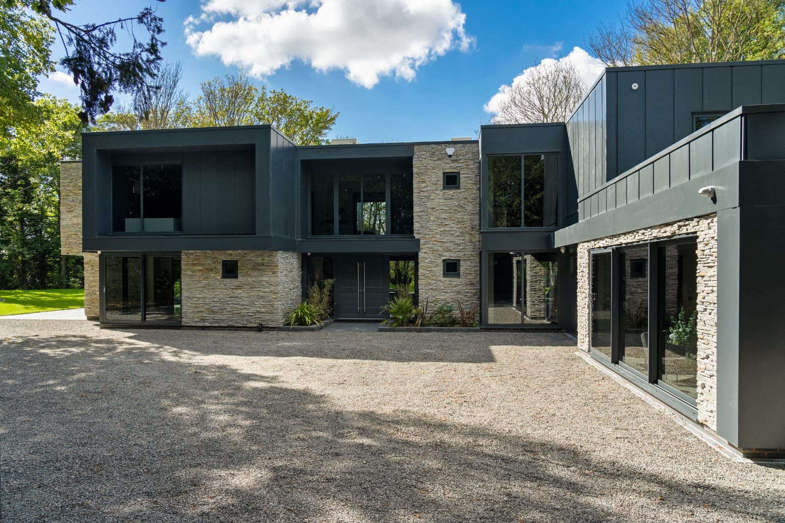 Nowoczesny dwupiętrowy dom z elewacją z kamienia i ciemnoszarego metalu, dużymi oknami i żwirowym podjazdem.