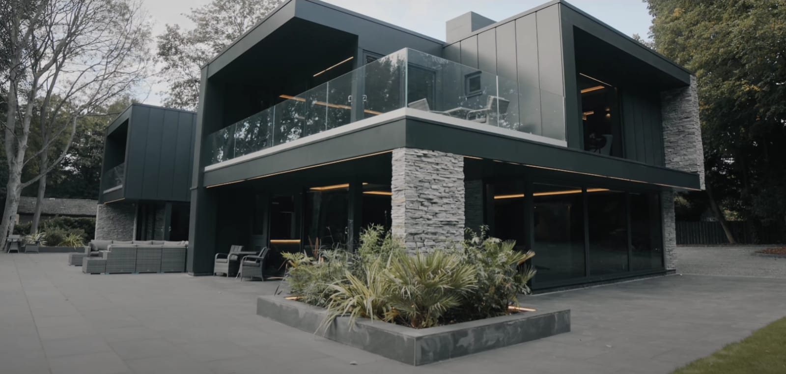 Nowoczesny, ciemnoszary dom z kamiennymi akcentami, szklanymi balustradami i betonowym patio. W tle drzewa.
