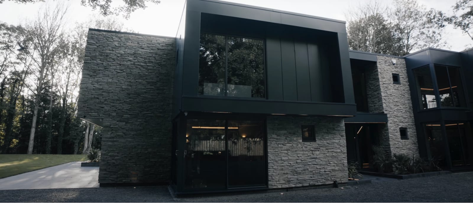 Nowoczesny, dwupiętrowy dom z kamienną i czarną fasadą. Otoczony drzewami, z podjazdem przed domem.