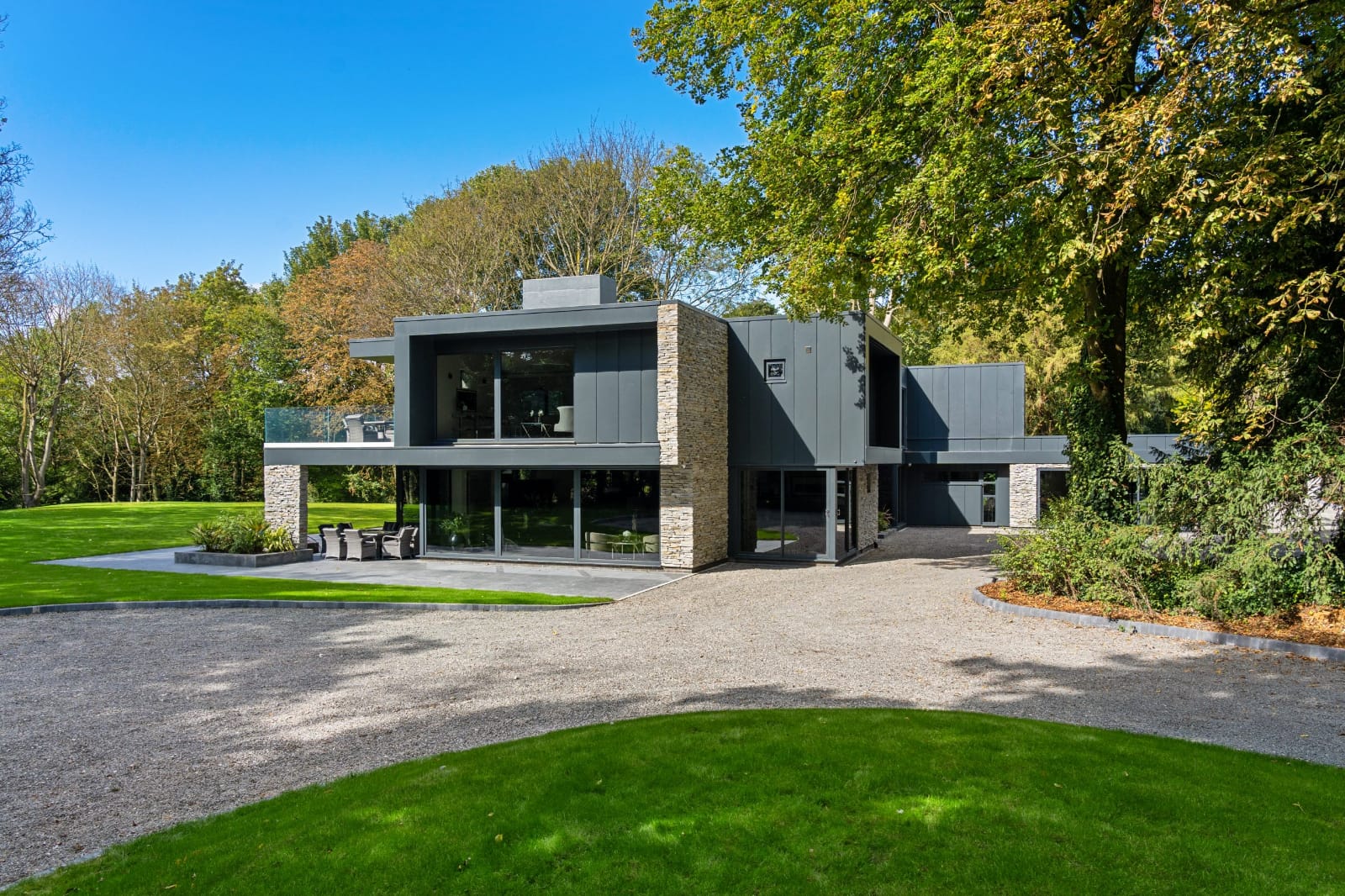 Nowoczesny szary i kamienny dom z dużymi oknami, położony na zielonym trawniku z żwirowym podjazdem, otoczony drzewami.