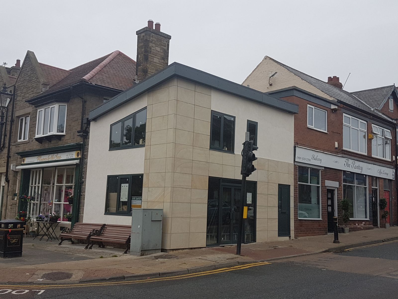 Dwupiętrowy budynek z beżową fasadą, czarnymi oknami i drzwiami, położony na rogu ulicy razem z innymi sklepami.