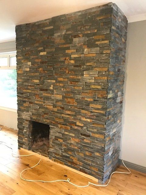 Kominek obłożony kamieniem, w odcieniach szarości i złota, wbudowany w ścianę. Biała listwa przypodłogowa i kable elektryczne na drewnianej podłodze.