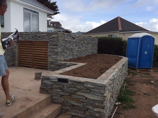 Mężczyzna nakładający uszczelniacz przy kamiennych murach i podwyższonej grządce ogrodowej z niebieską przenośną toaletą w tle.