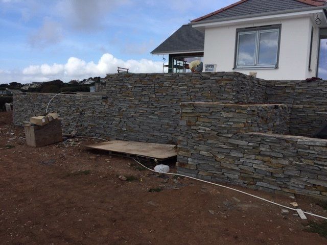 Kamienne mury oporowe wokół domu w budowie; brązowa ziemia, biały dom, pochmurne niebo.