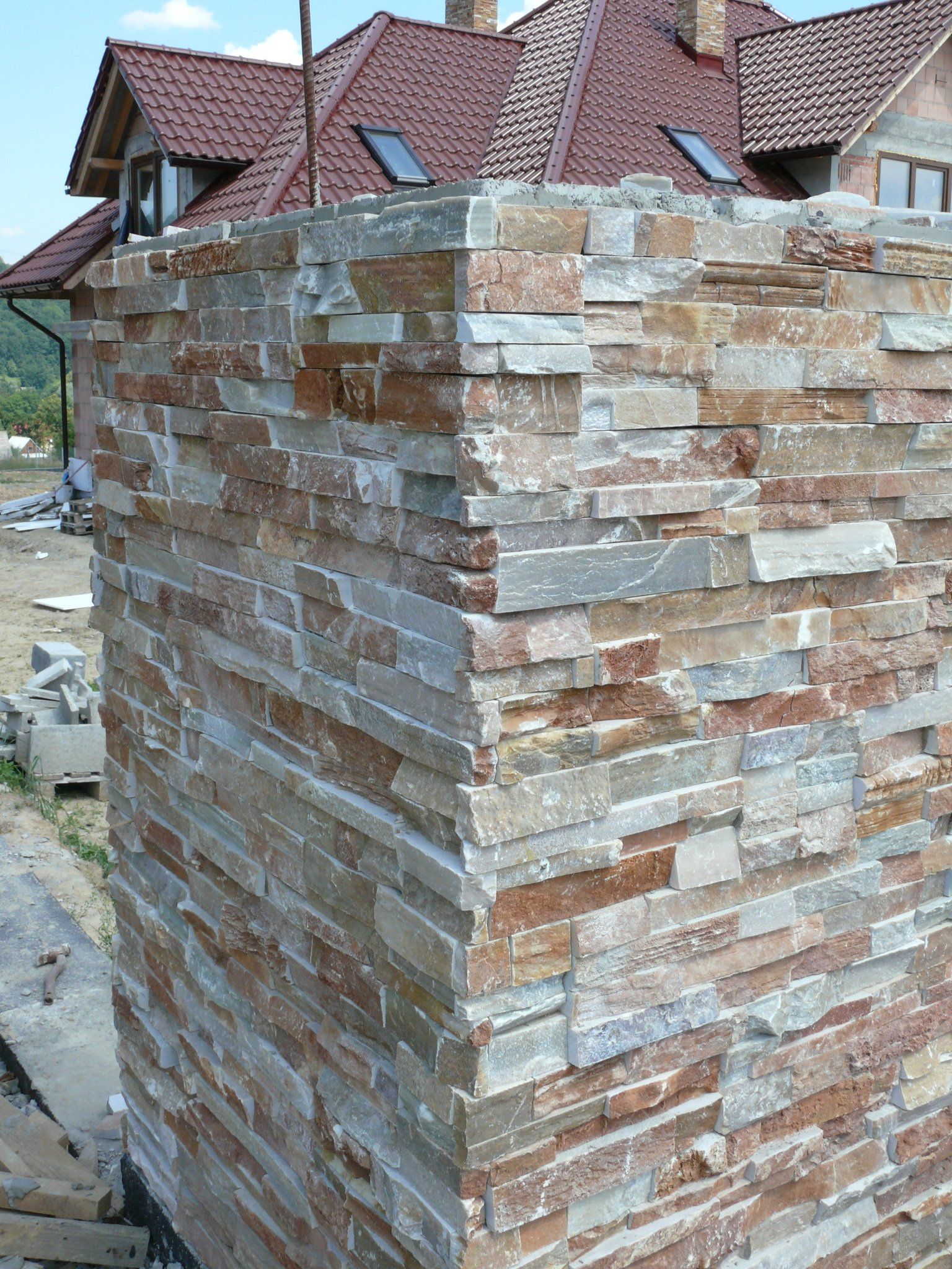 Narożnik budynku wyłożony kamieniem, z wielobarwnymi poziomymi pasami kamienia. W tle widoczny dach z czerwonymi dachówkami.