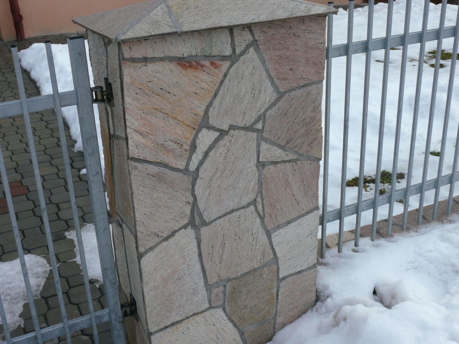 Kamienny słup z metalową bramą i ośnieżonym podłożem. Słup ma zwieńczenie.
