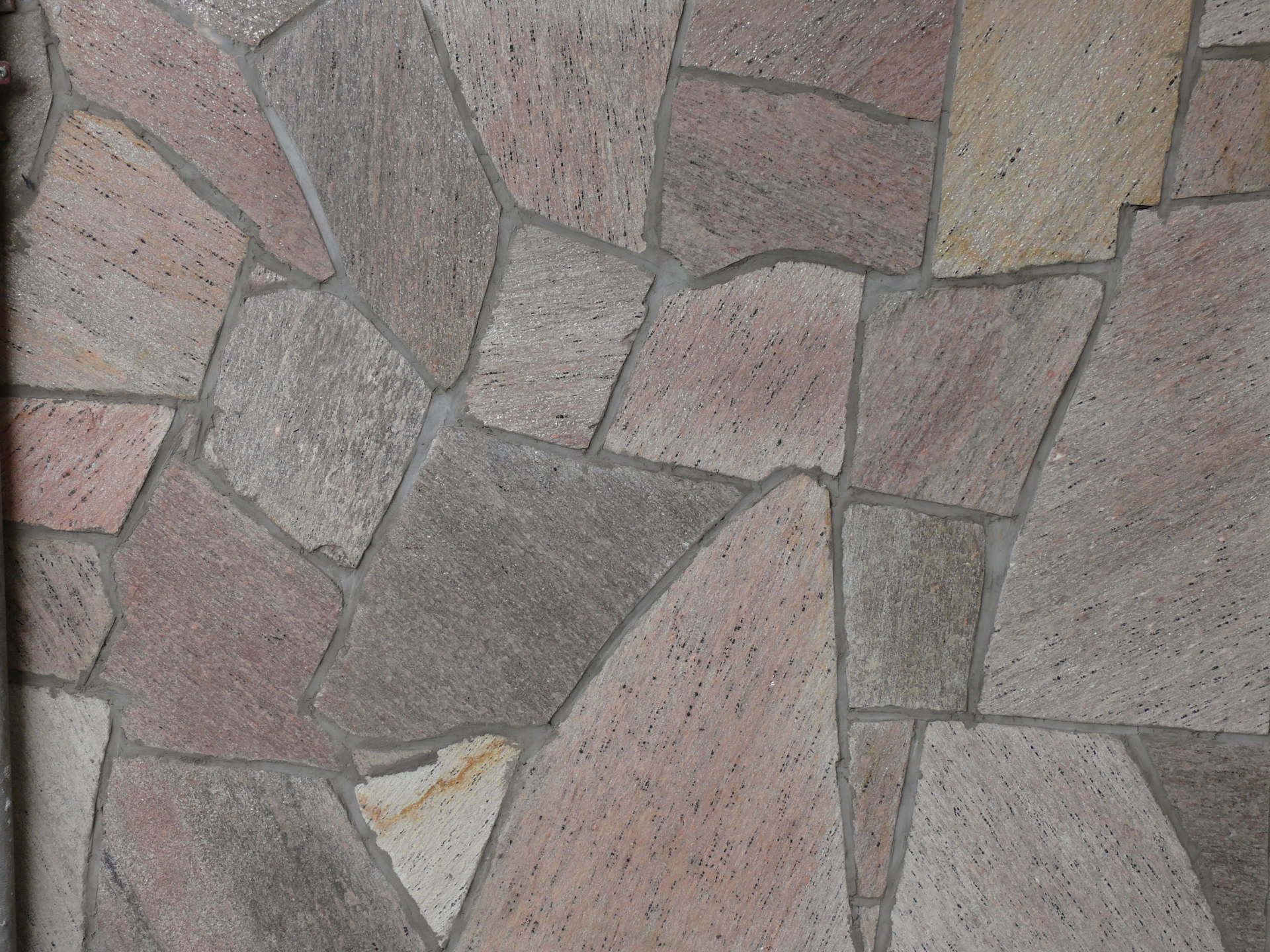 Mur kamienny, składający się z nieregularnych, wielokolorowych kamieni z szarą fugą.