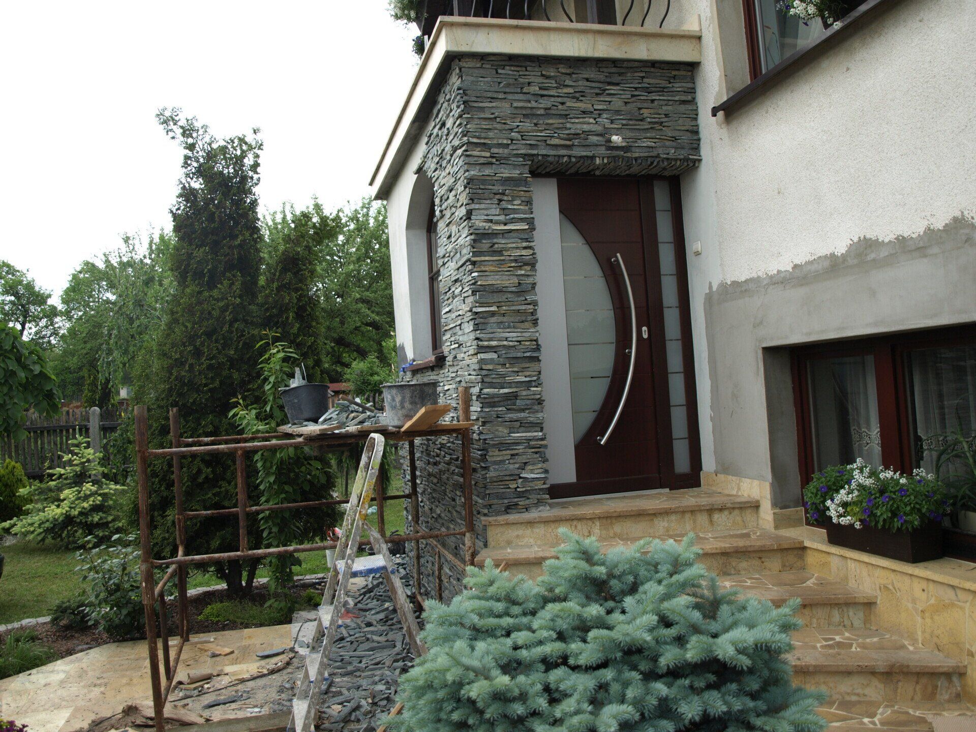 Wejście do domu z kamiennymi akcentami, brązowymi drzwiami i schodami. Na pierwszym planie mały krzew.
