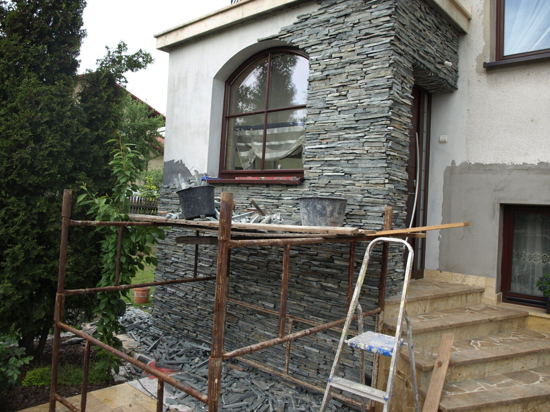 Elewacja domu w trakcie remontu z okładziną kamienną. Rusztowanie, narzędzia i częściowo wykończona ściana.