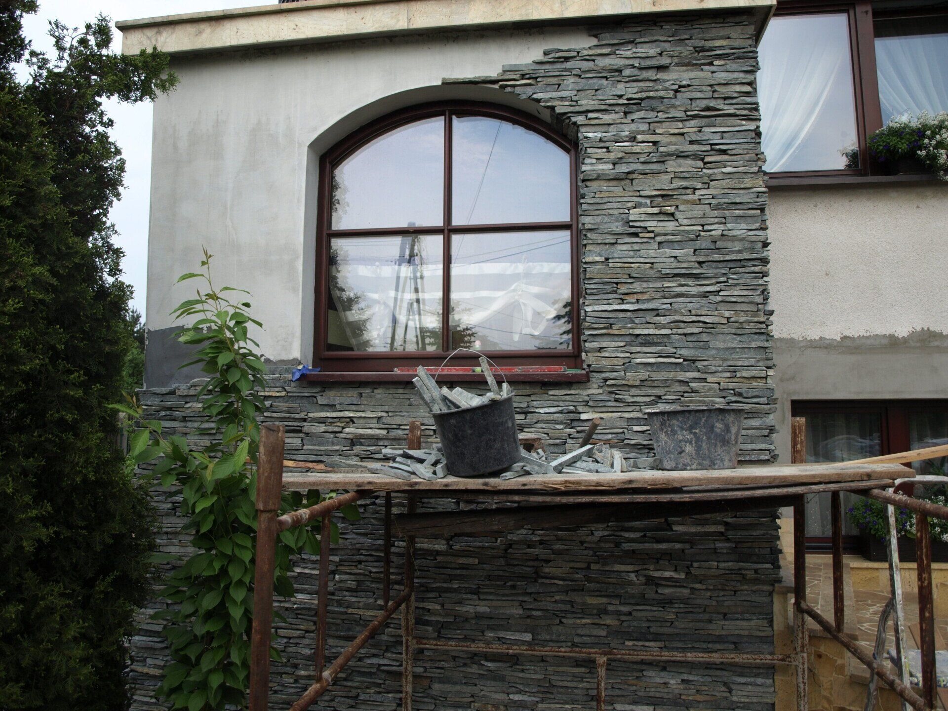 Zewnętrzna część budynku z łukowym oknem, częściowo obłożona kamieniem. Rusztowanie, wiadro i konstrukcja.