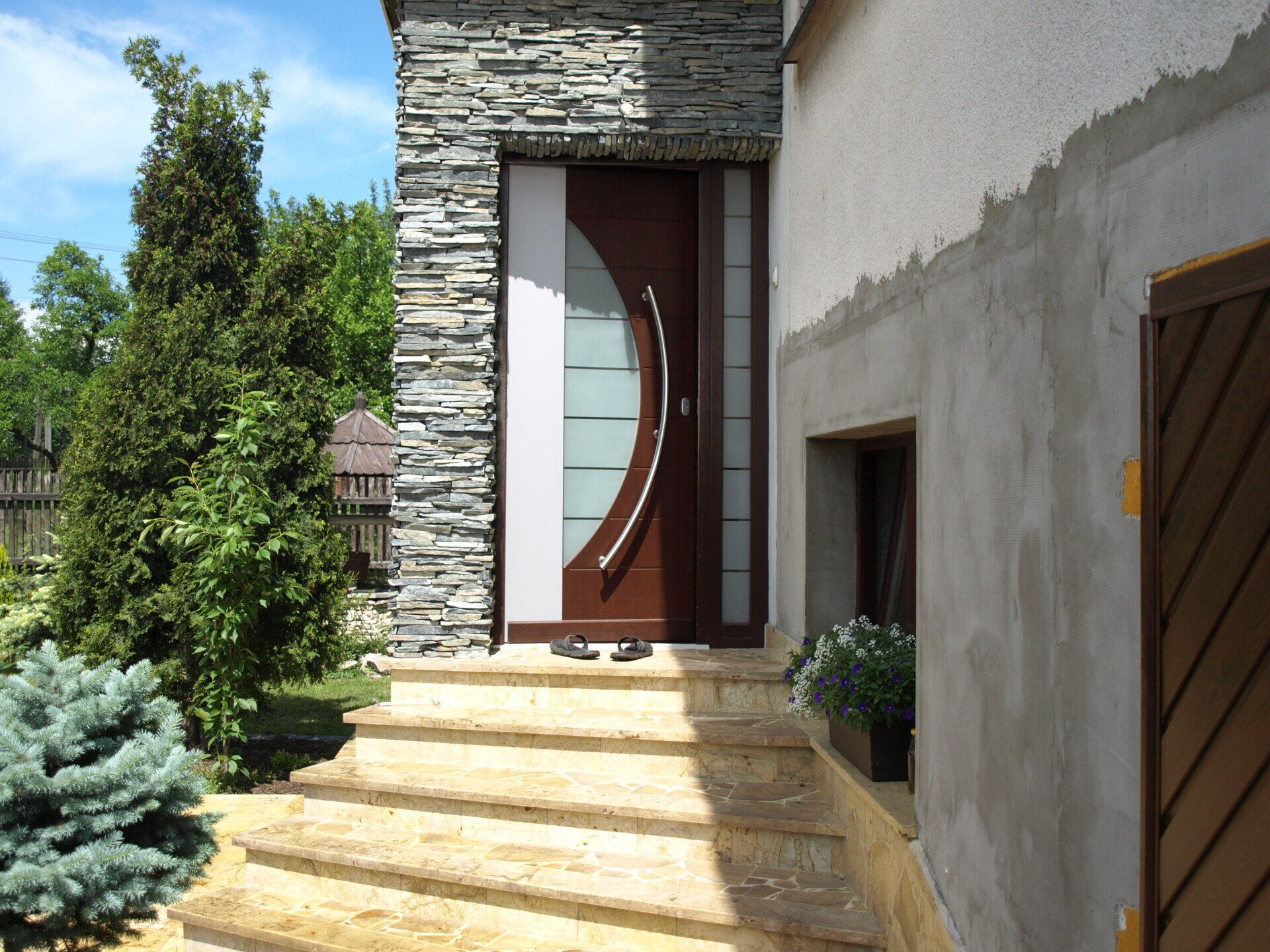 Brązowe drzwi wejściowe ze szklanym panelem, kamienna ściana i schody prowadzące do wejścia.