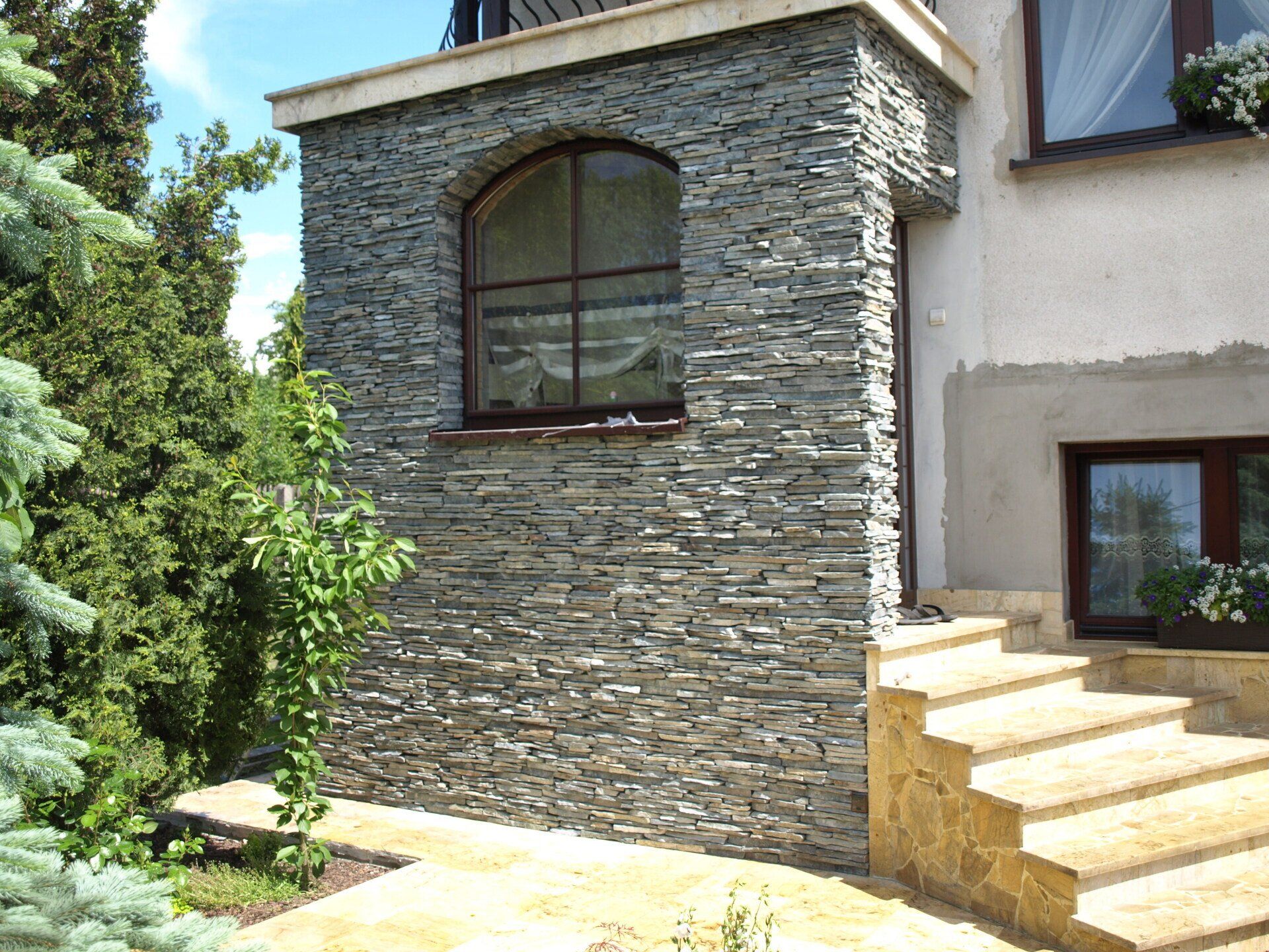 Kamienna ściana zewnętrzna z łukowym oknem, schodami i roślinami.