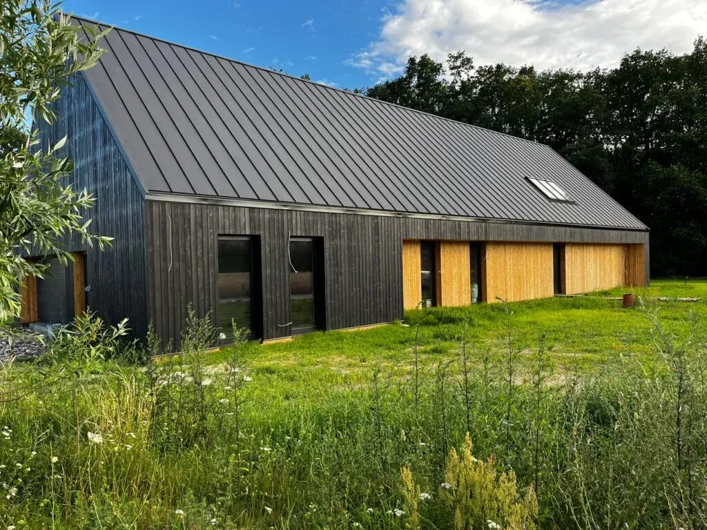 Nowoczesny dom z ciemnoszarym metalowym dachem i opaloną drewnianą elewacją, otoczony wysoką trawą.