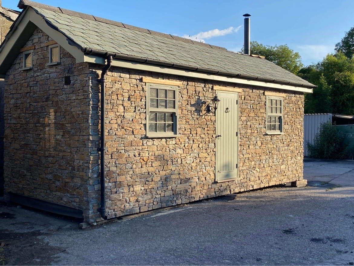 Kamienny domek z oliwkowozielonymi drzwiami i oknami; szary dach i komin.