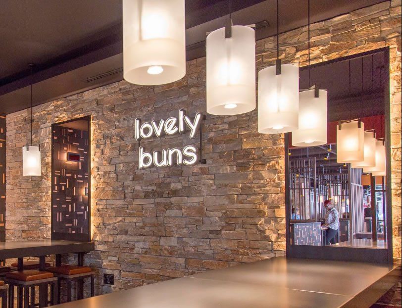 Wnętrze restauracji z szyldem „lovely buns”, kamienną ścianą, wiszącymi lampami i miejscami do siedzenia przy barze.