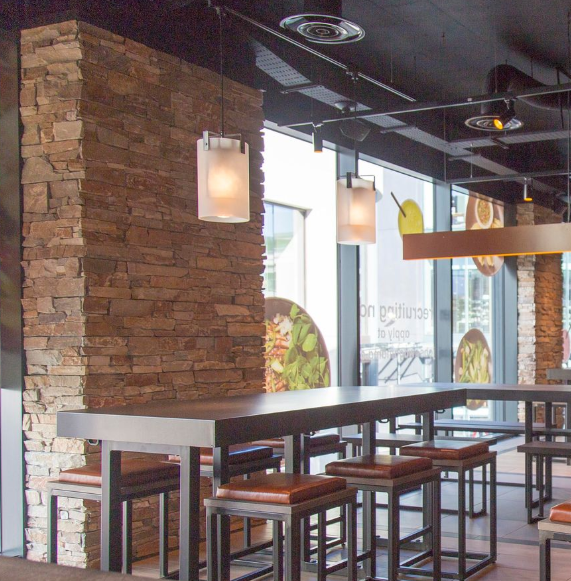 Wnętrze restauracji z kamienną kolumną, długim, ciemnym stołem ze stołkami i matowymi lampami wiszącymi.