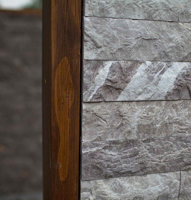 Drewniany słupek obok ściany z płytek kamiennych. Ciemnobrązowy słupek z jasnobrązową smugą. Szare płytki kamienne.