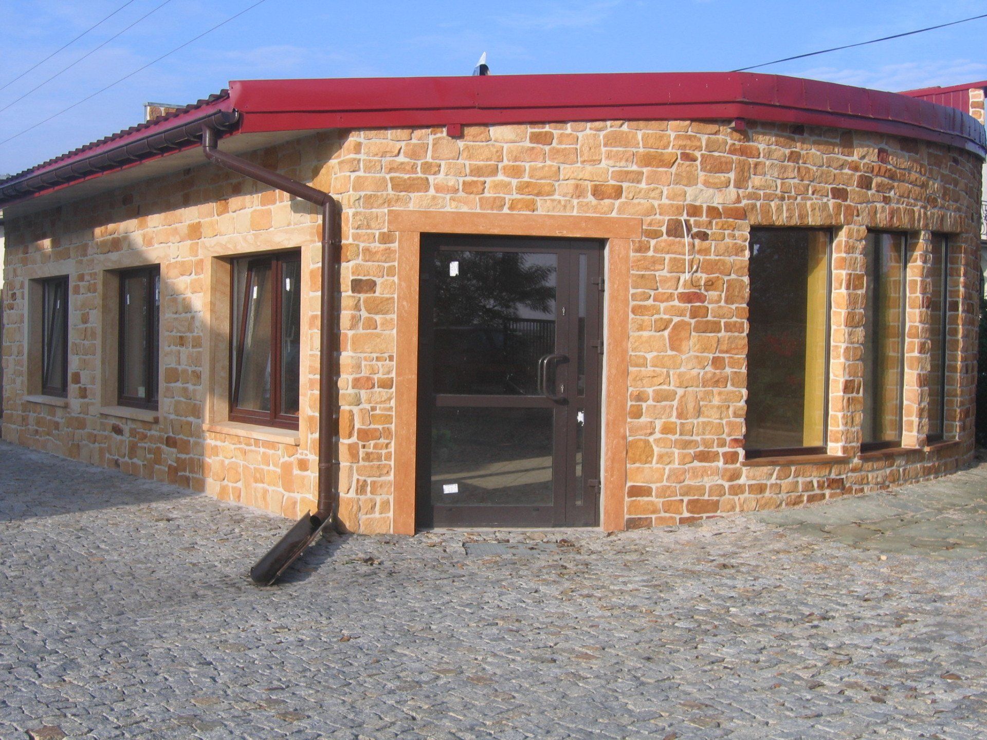 Ceglany budynek z czerwonym dachem oraz brązowymi ramami okien i drzwi.