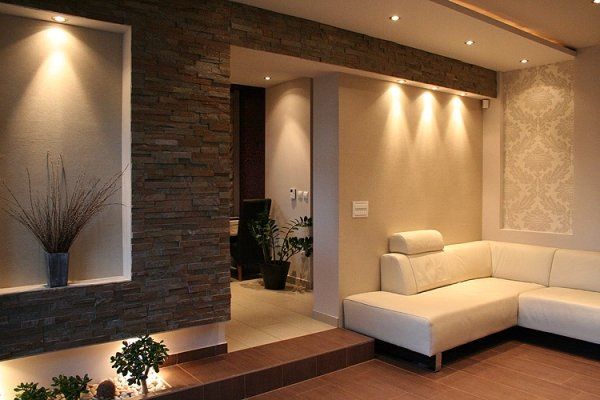 Nowoczesny salon z neutralnymi ścianami, oświetleniem wpuszczanym, kamienną ścianą akcentową i białą, narożną sofą.