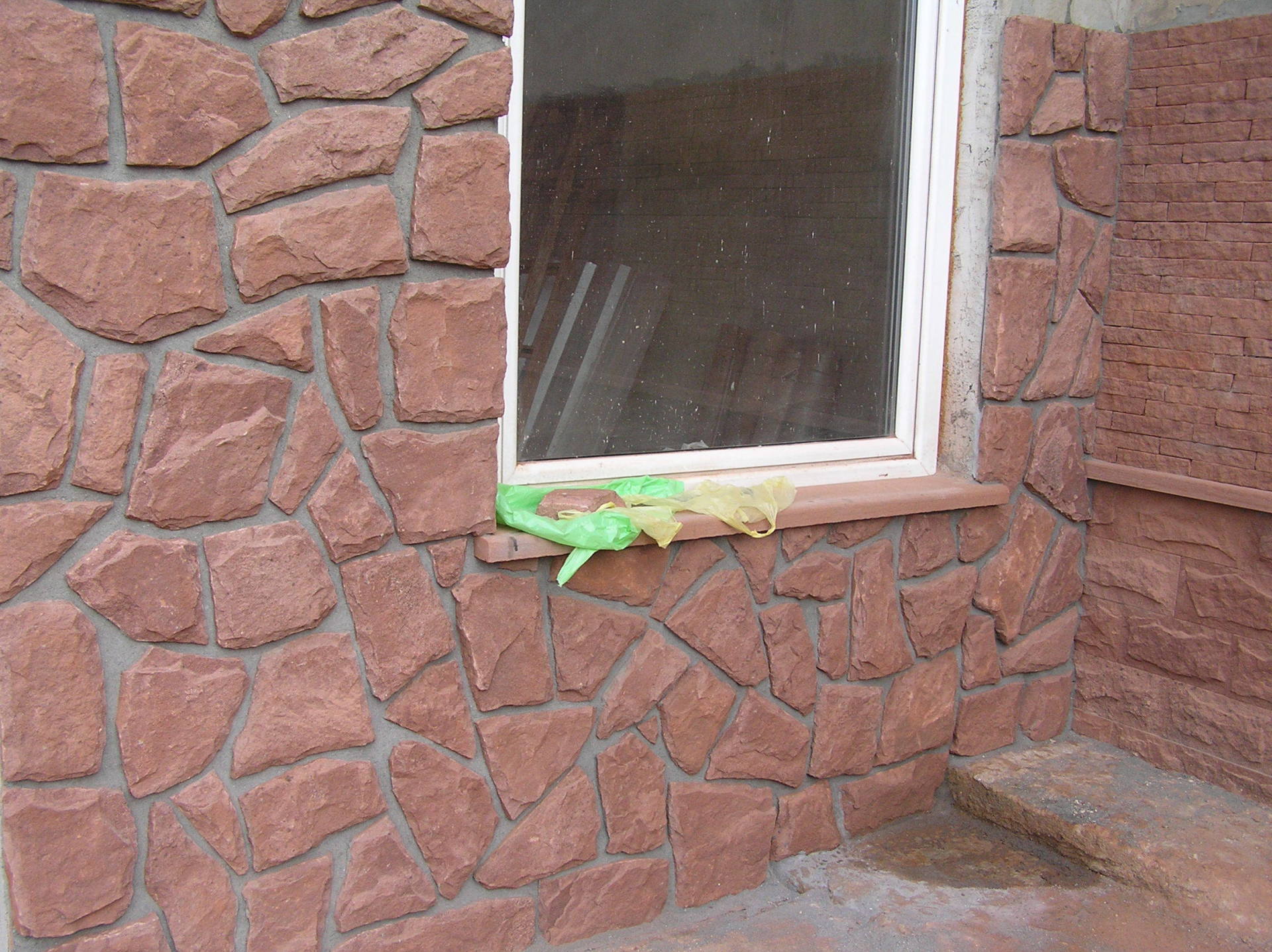 Kamienna fasada z oknem, z czerwonobrązowymi kamieniami i białą ramą okienną. Zielono-żółty plastik na gzymsie.