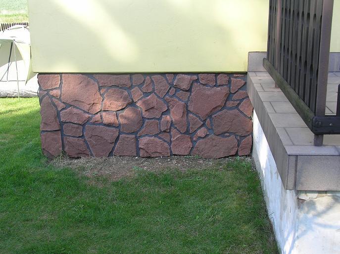 Kamienny fundament budynku; brązowe kamienie z ciemną zaprawą na tle żółtej ściany i zielonej trawy.