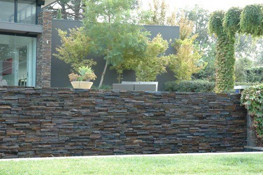 Ciemny kamienny mur oporowy przed nowoczesnym domem, w tle drzewa i trawa.