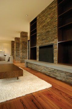 Salon z kamiennym kominkiem, wbudowanymi półkami, drewnianą podłogą i białym dywanem.