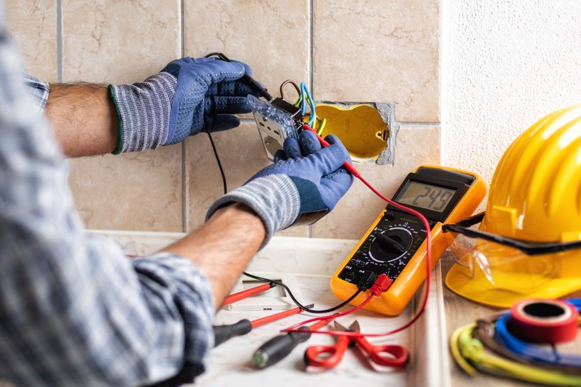 An electrician wearing gloves uses a multimeter to test wires in a wall outlet, with tools and a yellow hard hat nearby.