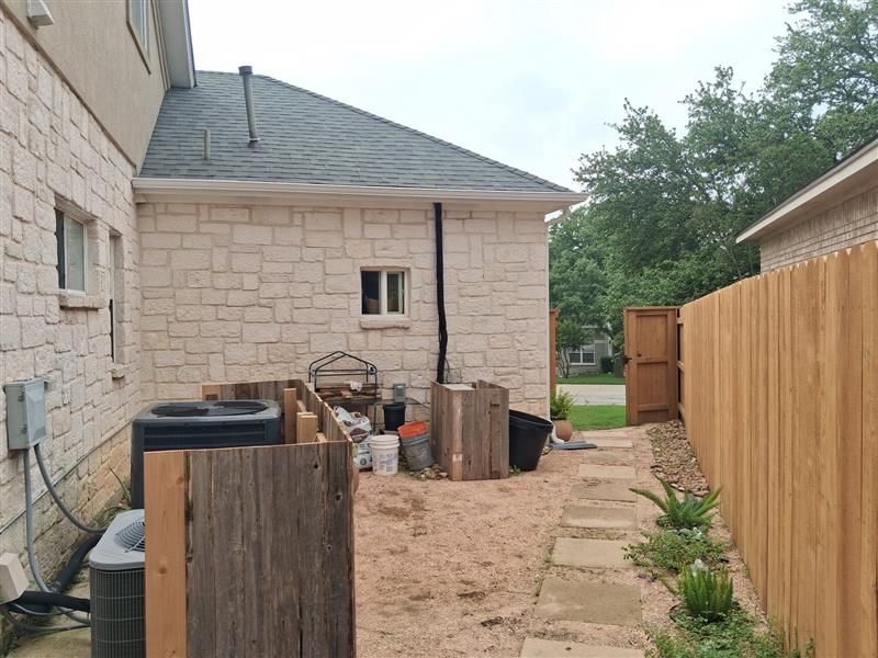 Narrow side yard with stone house, wooden fence, stepping stones, and stacked outdoor items on sandy ground