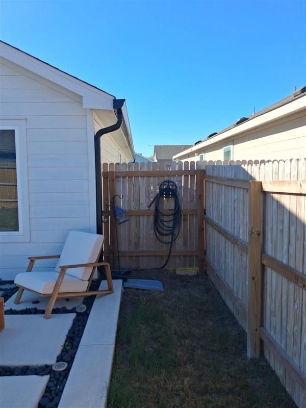 Narrow backyard walkway beside a house, with a fence, hose, and two white chairs on a patio.