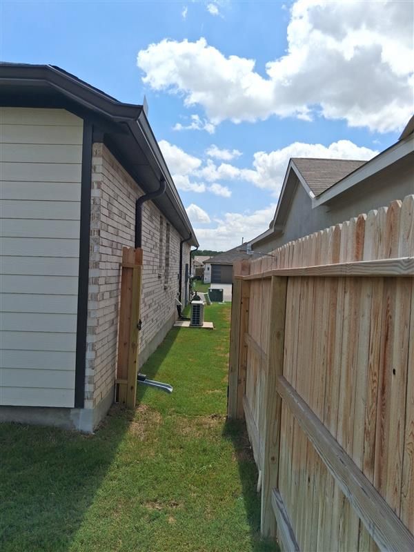 Narrow grass alley between a house and wooden fence under a blue sky with clouds.