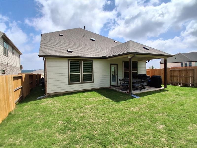 Backyard view of a beige house with a covered patio, fenced lawn, and partly cloudy sky
