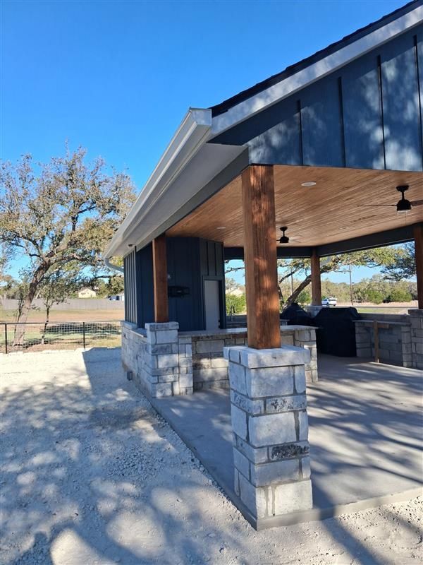 Covered outdoor patio with stone columns and a kitchen area, beside a shaded building under a blue sky