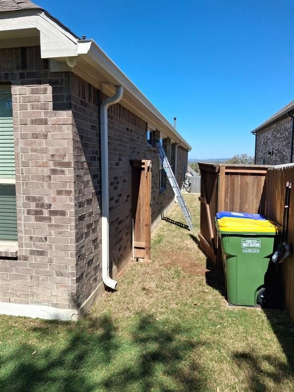 Side of a brick house with white gutters, narrow grass strip, and two trash bins near a wooden fence.