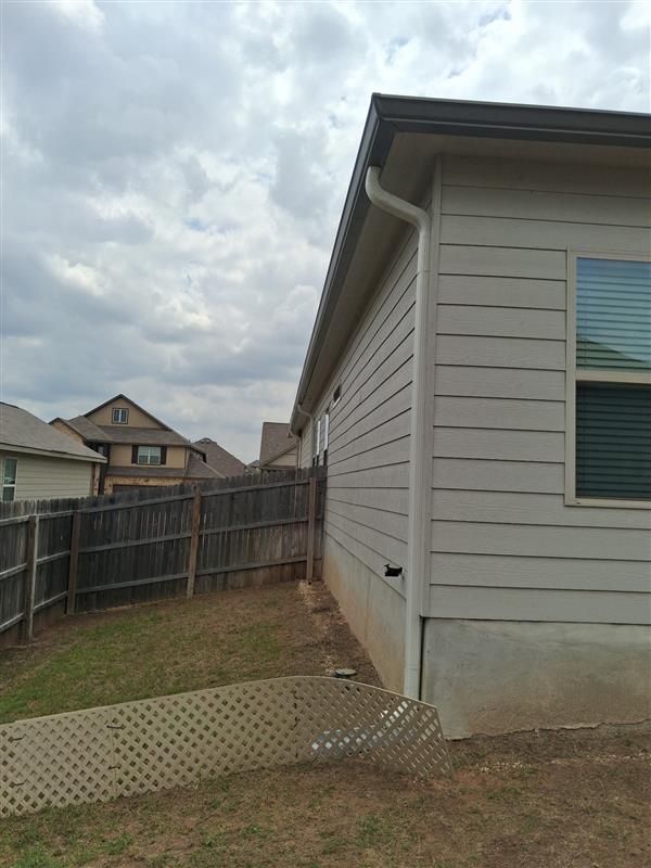 Side yard with beige house siding, wooden fence, concrete slab, and cloudy sky.