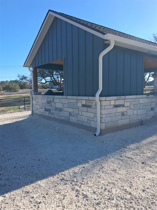 Blue-sided pavilion with stone base and white gutter, standing on a gravel lot under a clear sky