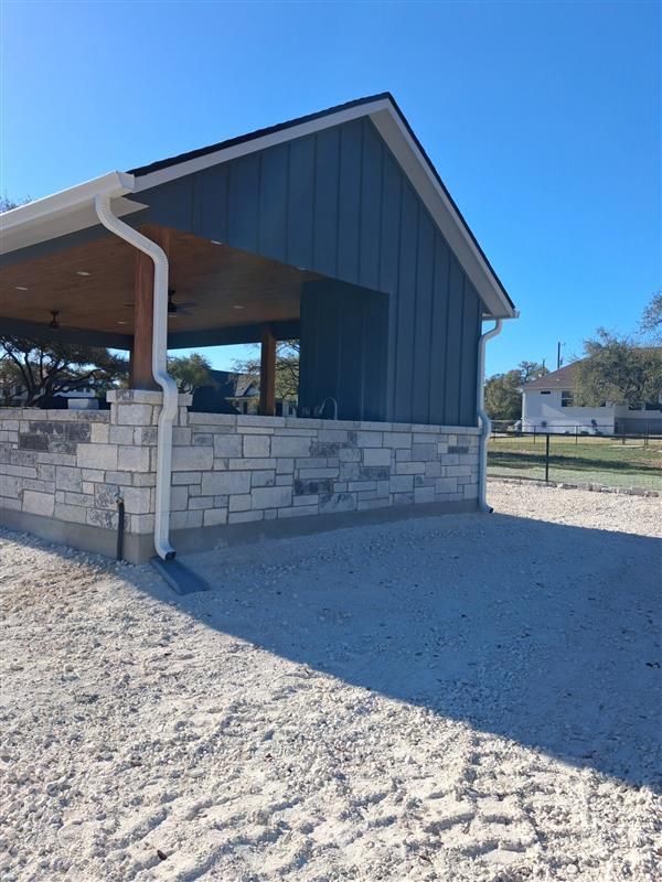 Stone pavilion with dark blue siding and a covered porch on a gravel lot under a clear blue sky