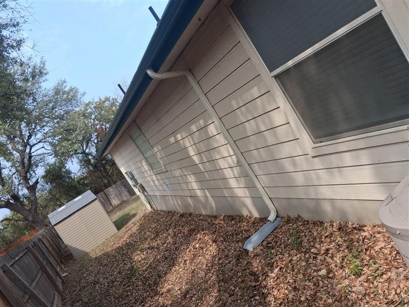 Side yard of a house with tan siding, roof eaves, a downspout, and leaf-covered ground beside a wooden fence.