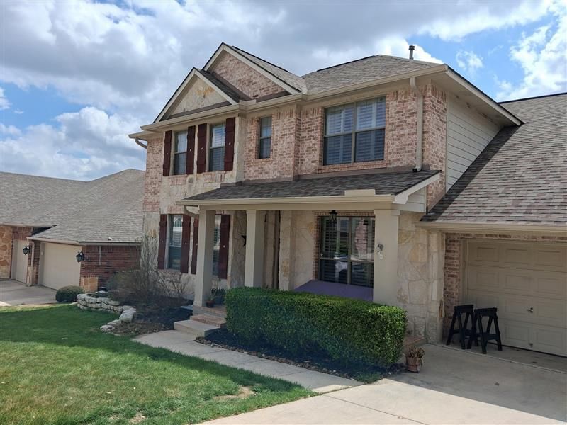 Two-story brick suburban house with a front porch, manicured lawn, and driveway under a cloudy sky