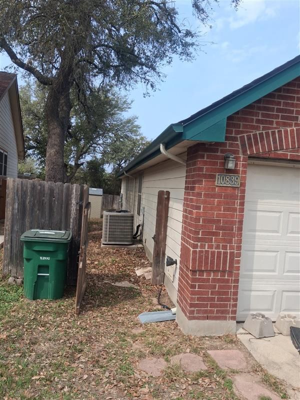Backyard side view of a brick house with a garage, green trash bin, and air conditioner unit under trees