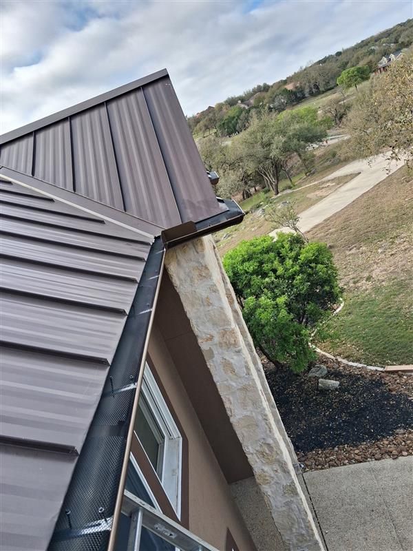 Tilted view of a house roof with dark metal panels, gutter, and a hillside yard below