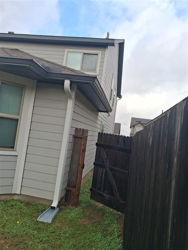 Side of a gray house with a white downspout beside a dark wooden fence and a grassy yard.