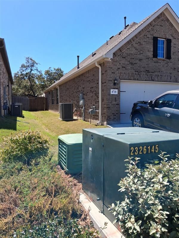 Brick house side yard with utility boxes, driveway, and parked car in bright sunlight