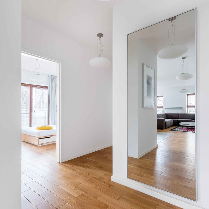 Couloir lumineux avec parquet, grand miroir et aperçu d'une chambre blanche avec un lit et des suspensions lumineuses.
