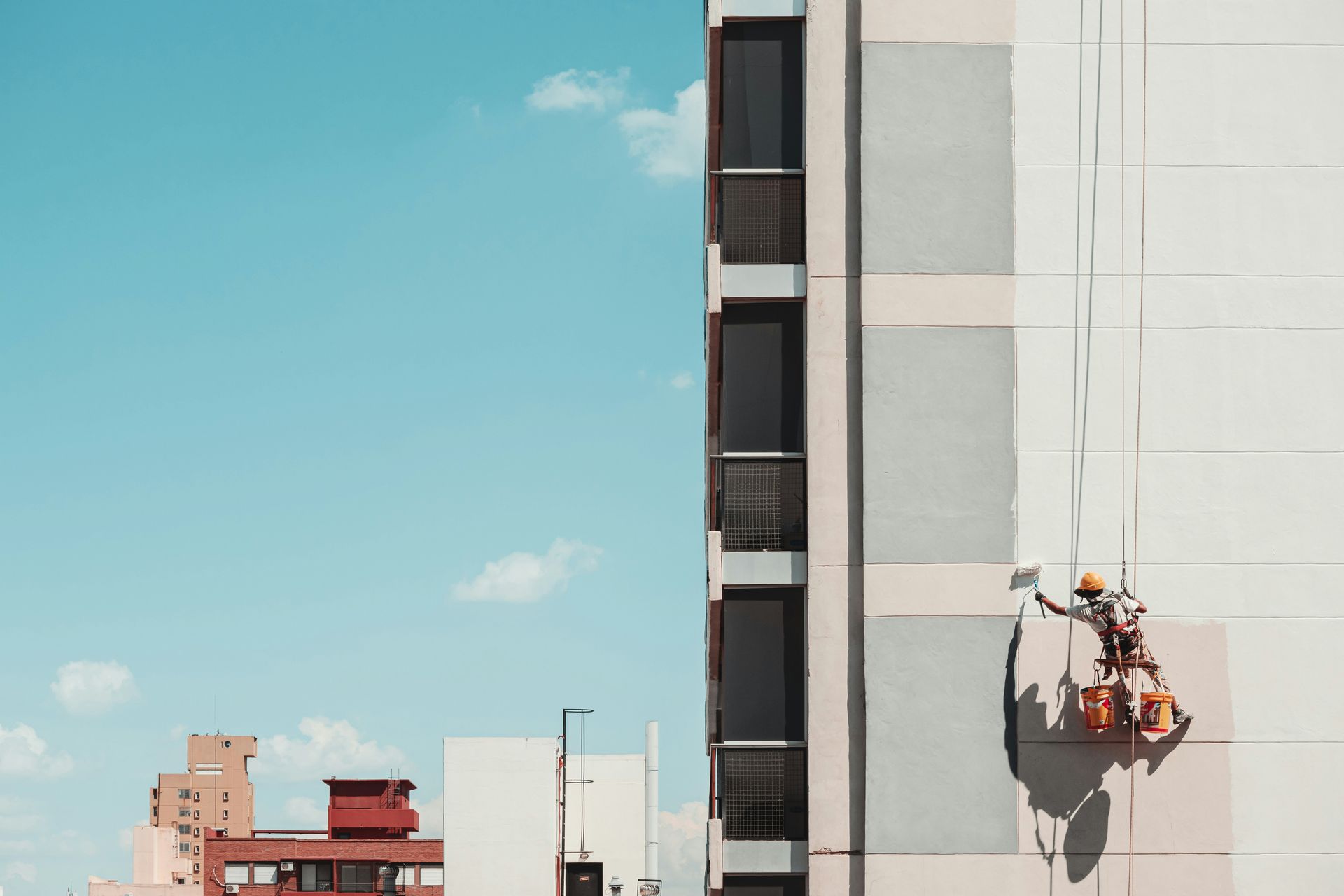 A worker in a red scissor lift uses a long-handled roller to paint a smooth, light-colored wall against a clear blue sky.