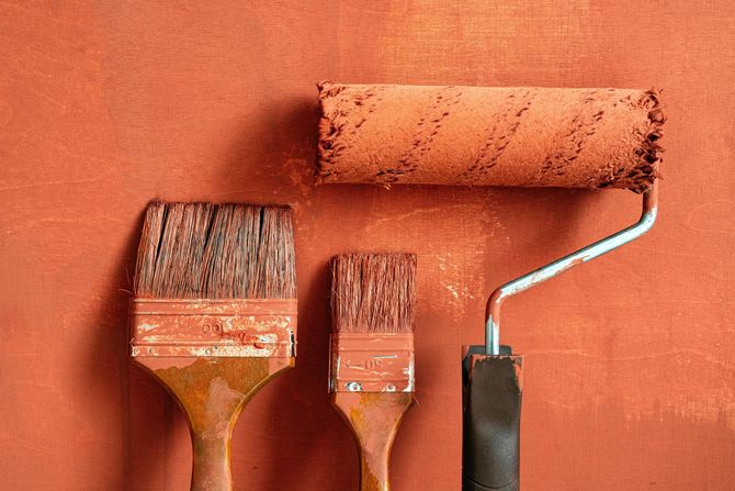 A paint roller and two paintbrushes covered in terracotta-colored paint, resting against a painted wall of the same color.